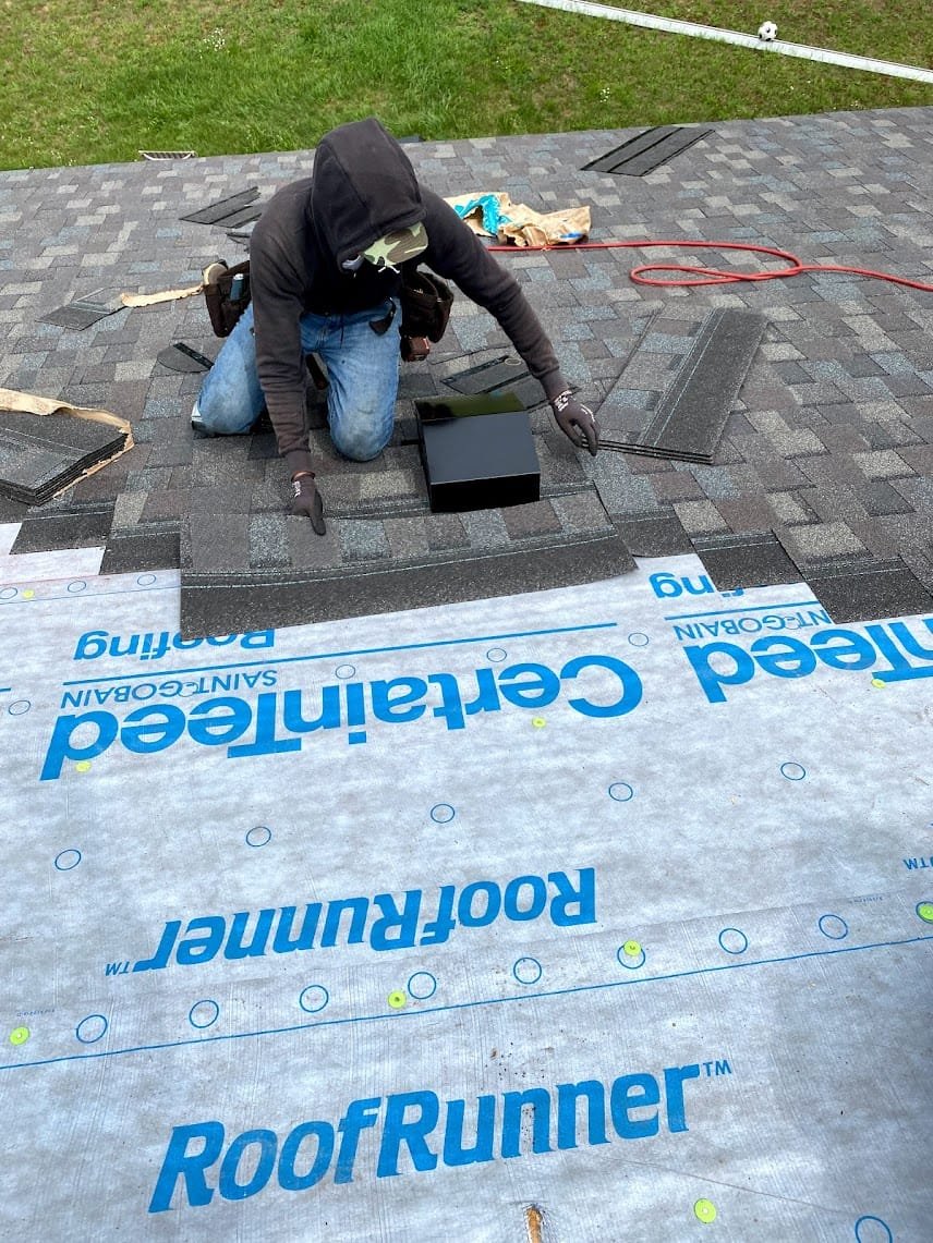 Person installing shingles on a roof with underlayment labeled "RoofRunner" visible, wearing a hoodie and gloves.