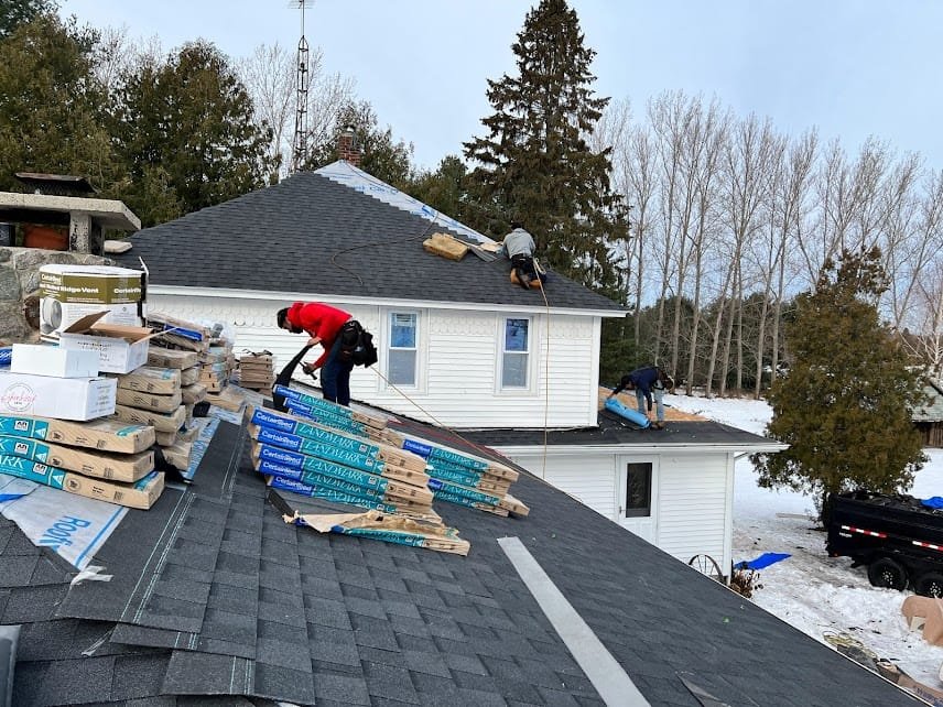 Workers install shingles on a snowy rooftop, surrounded by roofing materials and dense trees in the background.