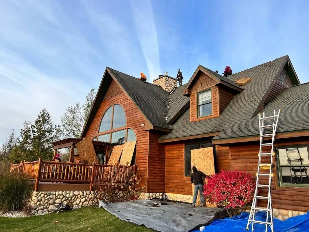 Workers are installing boards on the roof of a two-story wooden house with a ladder on the side and a tarp on the ground.