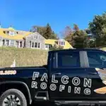 A black Falcon Roofing truck is parked on a road, with a large house featuring a yellow roof in the background.