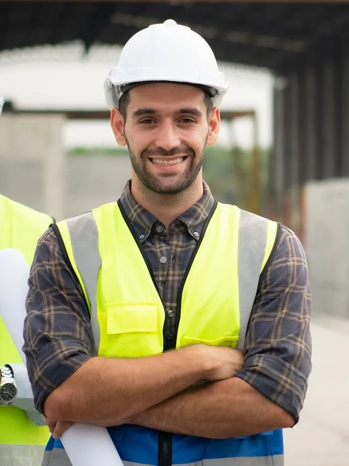A man wearing a white hard hat, plaid shirt, and yellow safety vest stands outdoors, holding rolled-up papers and smiling.