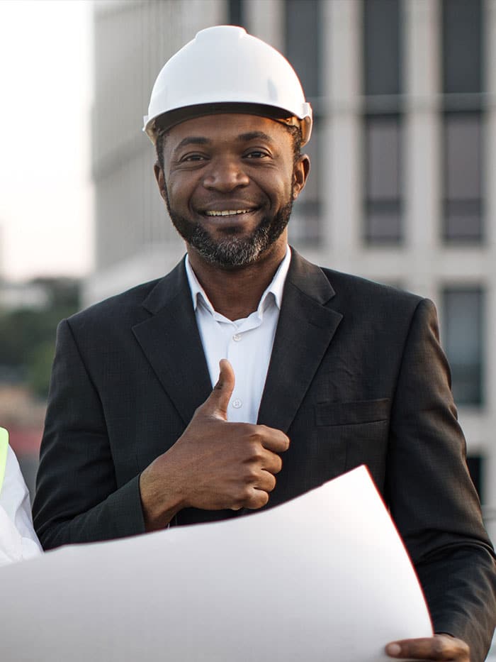 A man in a suit and hard hat smiles and gives a thumbs-up while holding architectural plans outdoors.