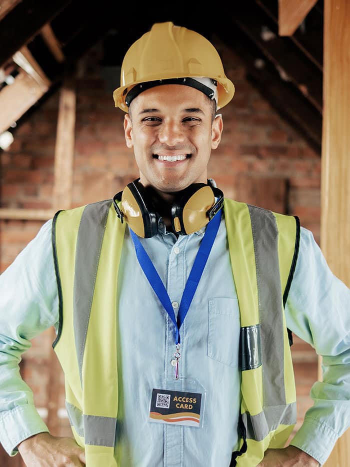 Smiling worker wearing a hard hat, safety vest, and headphones, with an access card around the neck, standing in a partially constructed building.