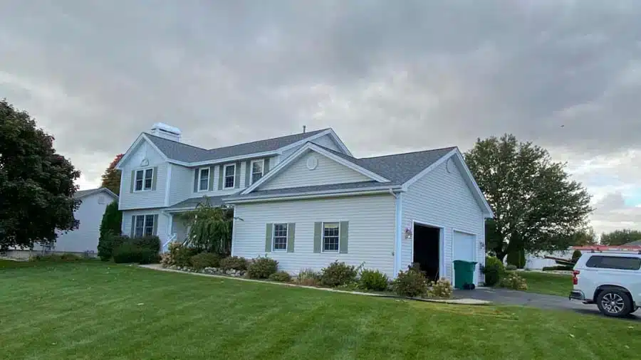 A two-story white house with a gray roof sits on a green lawn under a cloudy sky. A driveway leads to the garage, with a green bin and a vehicle nearby.