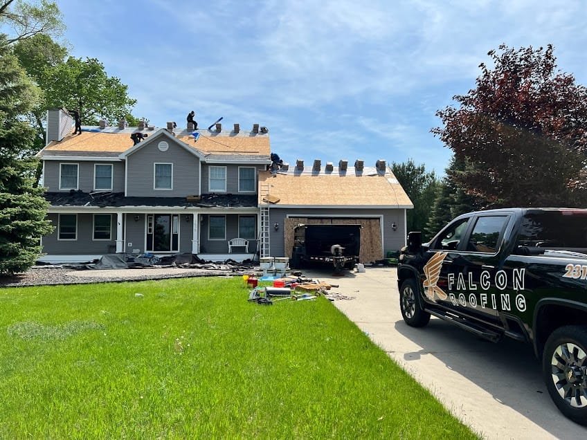 Workers replacing the roof of a two-story house, with bundles of shingles lined along the roof. A Falcon Roofing truck is parked in the driveway.