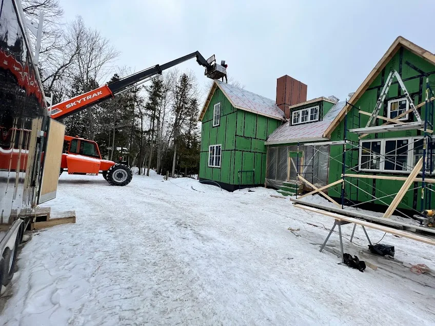 A construction site with a partially built green house. A forklift is lifting materials to the roof. Snow covers the ground.