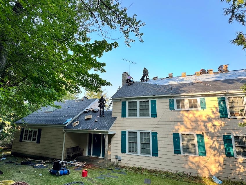 Workers repairing the roof of a two-story house with green shutters. Supplies and tools are scattered on the ground and roof. Trees surround the house under a clear blue sky.