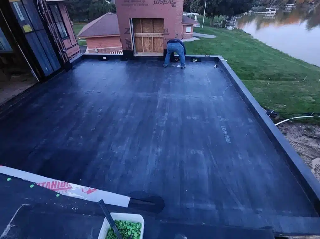 A worker expertly installs a flat roofing membrane on a residential building near a lake, where the lush greenery adds tranquility to the scene.