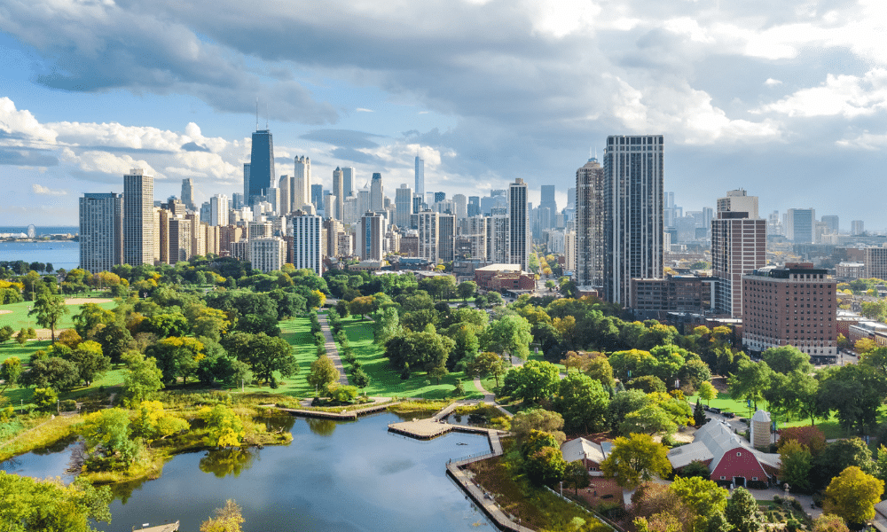 Aerial view of Chicago skyline with tall skyscrapers. Green park and small lake with foliage in the foreground under a partly cloudy sky.