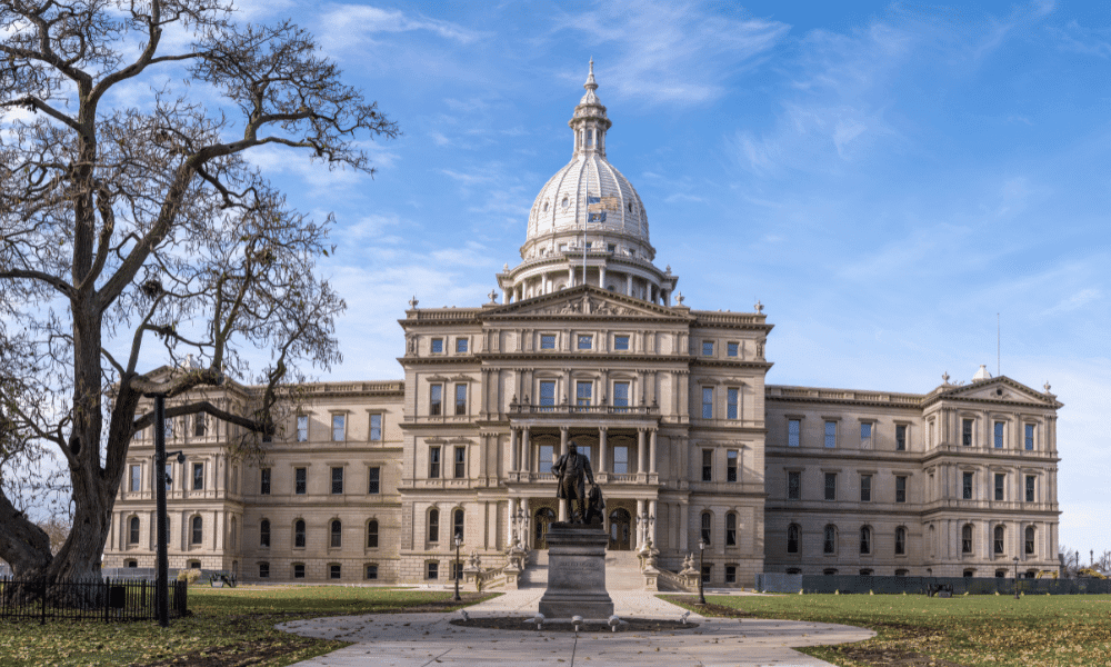 The image shows the Michigan State Capitol building, featuring a large dome, classical architecture, and a central statue in the foreground. A tree is on the left under a blue sky.