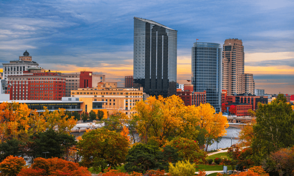 City skyline with tall buildings, trees with autumn foliage in the foreground, and a river running through the scene under a cloudy, colorful sky.