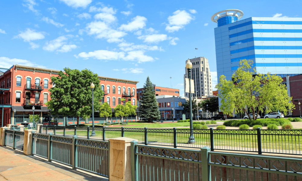 A small urban park is bordered by a green fence, with trees, red brick buildings, and a modern blue glass building in the background under a blue sky with scattered clouds.