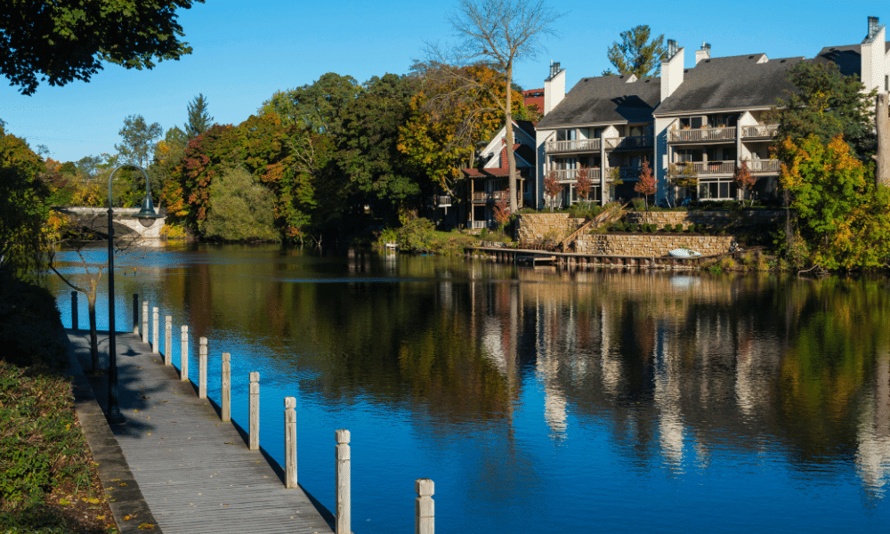 A serene river bordered by a wooden walkway and residential buildings with balconies. Lush trees with autumn foliage surround the area. A bridge is visible in the distance.