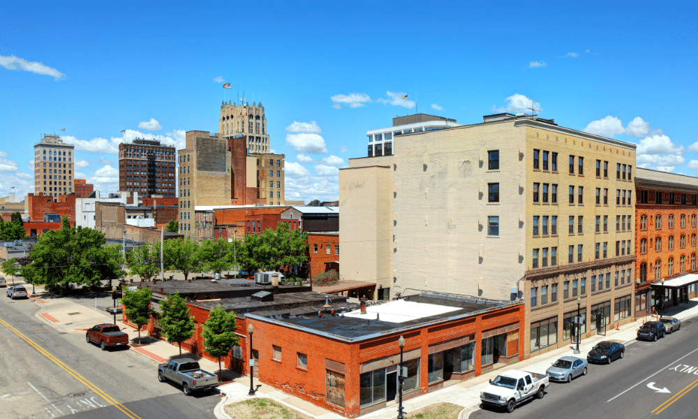 Cityscape with several multi-story buildings on a sunny day. Cars are parked along the street, and trees line the sidewalk. Skyscrapers are visible in the background against a clear blue sky.