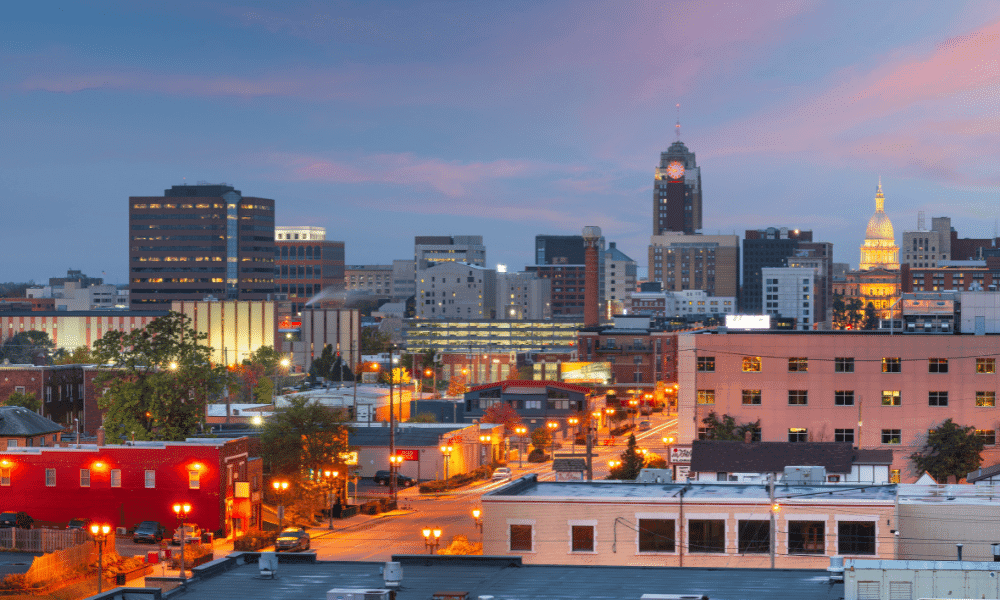 City skyline at dusk with a mix of modern and older buildings, lit streets, and a prominent domed structure in the background under a pastel sky.