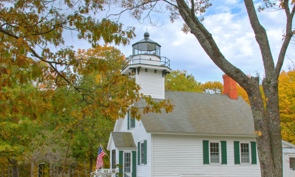 White lighthouse building with a chimney, surrounded by autumn trees. An American flag is displayed near the entrance. Sky is partly cloudy.