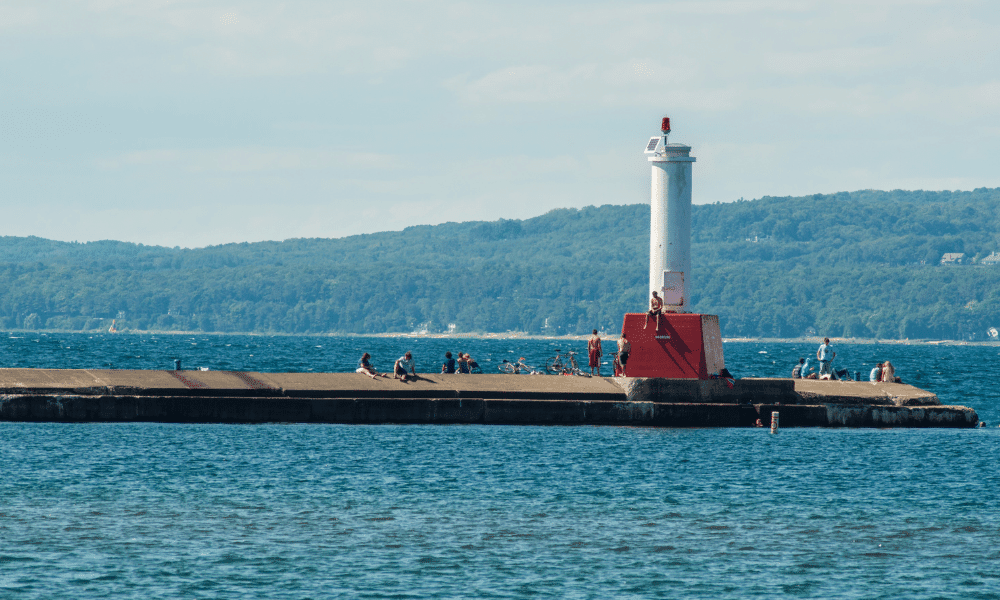 People sitting and standing on a pier with a white and red lighthouse, surrounded by water and distant green hills.