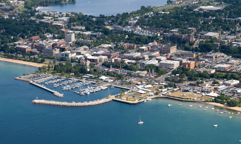 Aerial view of a coastal town with a marina, boats docked, and a small harbor. The town features numerous buildings, and a tree-lined landscape extends into the distance.