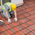 A worker in a high-visibility vest and hard hat repairs a red tiled roof with tools and sealant next to an open window, showcasing why professional roof inspections matter.