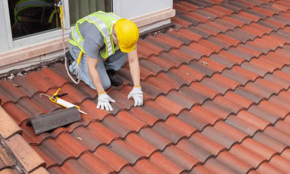 A worker in a high-visibility vest and hard hat repairs a red tiled roof with tools and sealant next to an open window, showcasing why professional roof inspections matter.