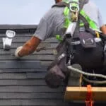 A person wearing safety gear and a tool belt works diligently, installing shingles on a roof. Standing on a wooden plank for support, they ensure every detail is perfected, avoiding overlooked roofing issues that could cost you thousands in the long run.