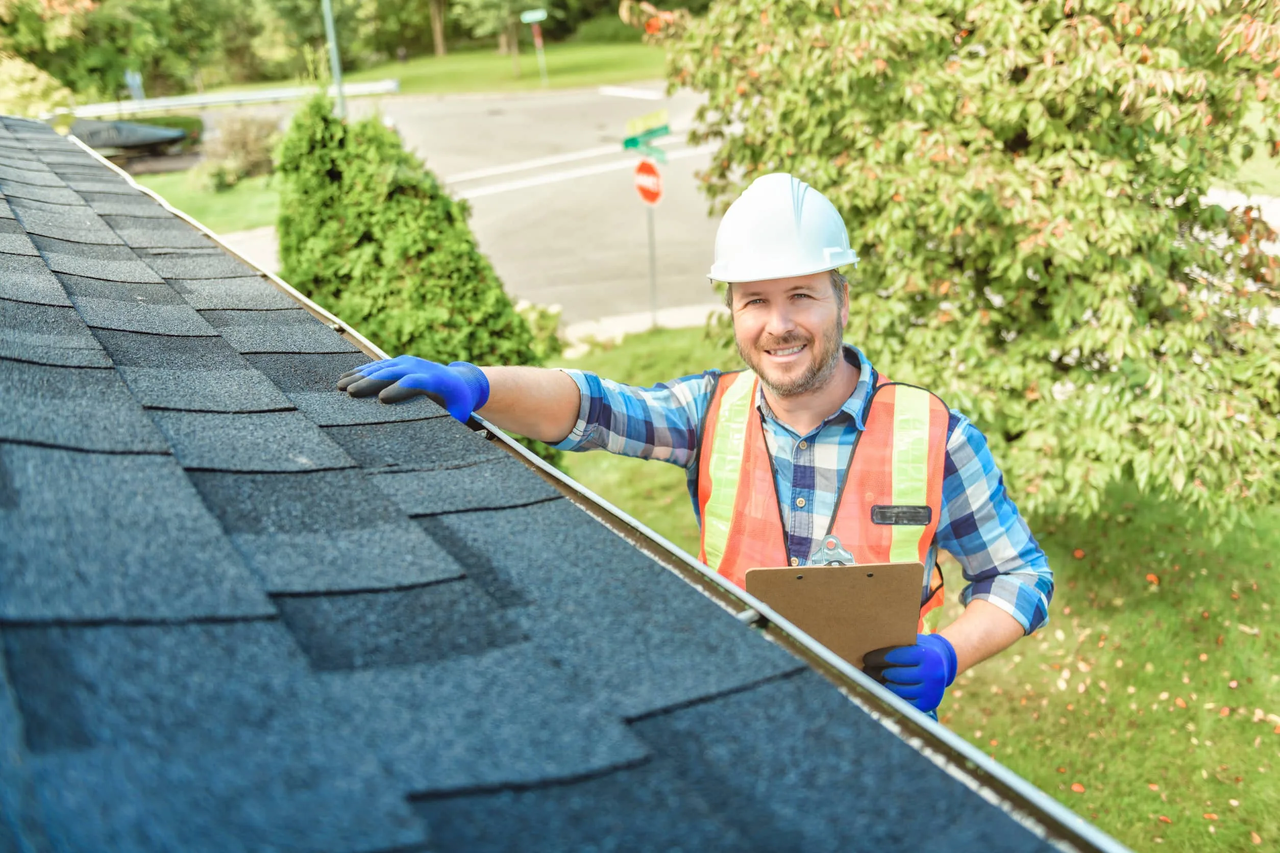 A construction worker, equipped with a hard hat and safety vest, diligently inspects a roof using a clipboard and gloves. It's part of the essential Seasonal Roofing Checklist, ensuring everything is in top condition on this sunny day outdoors.