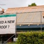A building under construction with scaffolding and a sign from local roofing companies reading "NEED A NEW ROOF?" displayed on the fencing.