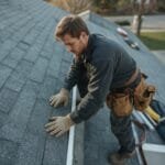A man wearing gloves and a tool belt installs or repairs shingles on a residential roof under daylight, demonstrating key steps from a homeowner guide to inspect your roof.