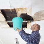 A man stands on a ladder holding a bucket under a leaking, damaged ceiling with exposed pipes and water stains, searching for roof leak solutions.