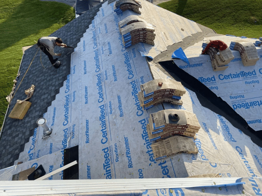 A person installs asphalt shingles on a sloped house roof, partially covered with CertainTeed roofing underlayment. Bundles of shingles, tools, and materials surround the site—a common scene for homes using professional roofing services.
