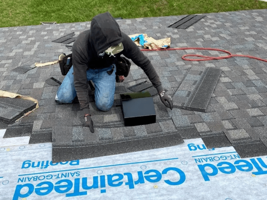 A person wearing a hooded sweatshirt and gloves installs asphalt shingles on a roof in Traverse City, kneeling by a vent opening. Roofing tools and materials are scattered nearby during the new roof installation on the partially shingled surface.